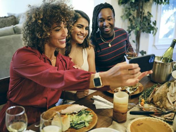 tre kvinner tar en selfie over et middagsbord med en bøtte med vin, kalkun, en gresskarpai, et stearinlys, tallerkener med mat på og et vinglass på bordet