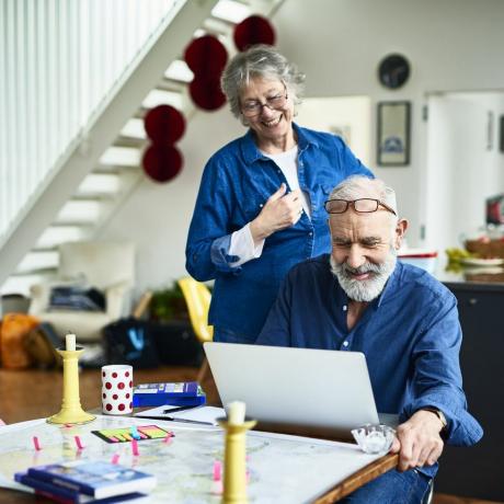 senior kvinne som smiler og ser over skulderen til mannen mens han undersøker og leser anmeldelser på nett om steder å bo, og steder å besøke, klistrelapper og guidebøker på bordet, booking av ferie på nett