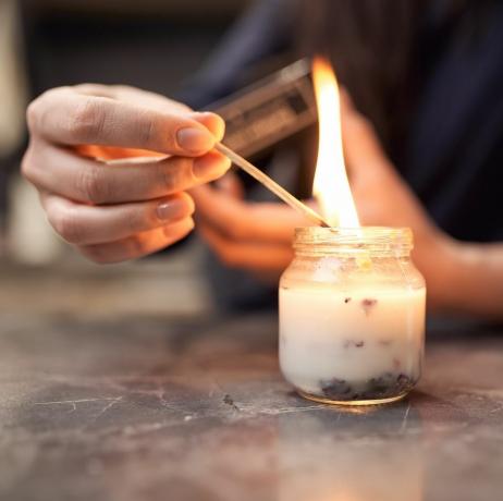 beskjær anonym kvinne med brennende fyrstikk som tenner aromatisk lys i glasskrukke plassert på marmorbord hjemme