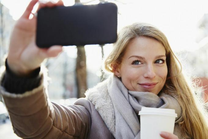 kjekk kvinne som går i byens gater om vinteren og tar en selfie og holder en kaffe for å gå, bakgrunnsbelyst