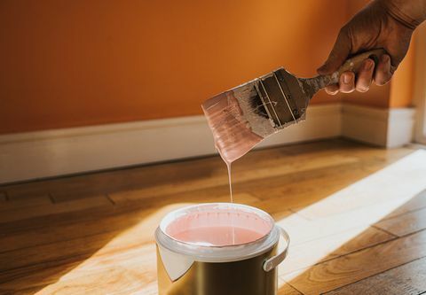 hånden holder en tykk pensel og fjerner den, etter å ha dyppet den i malingen lager den en dråpe maling som spruter tilbake i potten veggen bak er malt oransje og fargen i boksen er rosa, noe som kolliderer konseptuelt med plass til kopiere