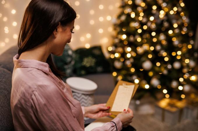 glad ung kvinne som sitter på sofaen, ved det glitrende juletreet, åpner og leser et julekort hun fikk med gaven