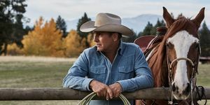 kevin costner i yellowstone ved siden av en hest som lener seg på et gjerde med tau i hendene iført en falmet blå denimskjorte og beige cowboyhatt
