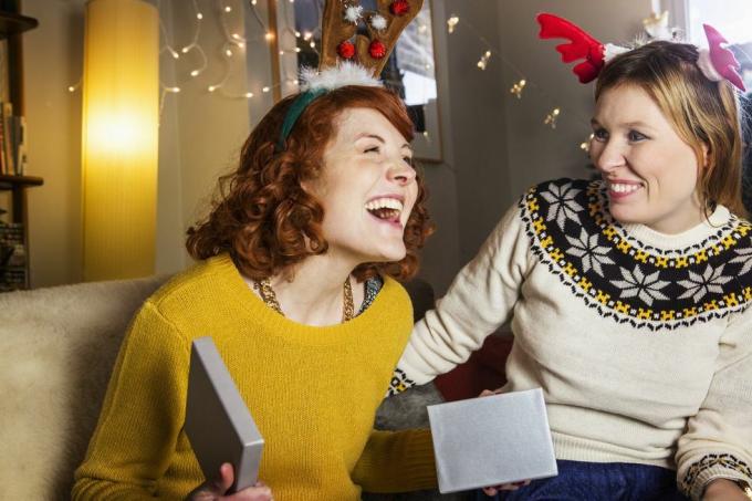 rødhåret kvinne smiler når hun sitter på en sofa og åpner en julegave i en sølvboks, det er en kvinne i en genser ved siden av henne og smiler hun har rosa og rødt gevir på toppen av hodet