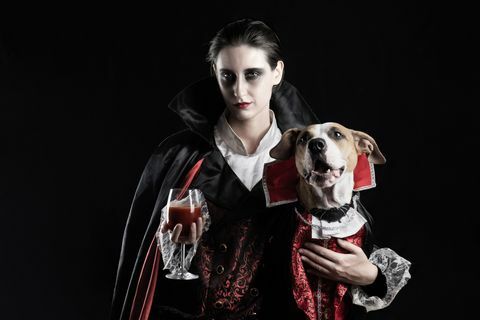ung kvinne med et glass rødt drikke og kjæledyrvalpen hennes kledd i samme dracula-kostyme