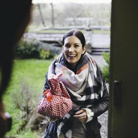en kvinne smiler mens hun står på dørtrinnet og besøker en venn hun er pakket inn i varme klær og holder en gave pakket inn i en rød og hvit rutete klut
