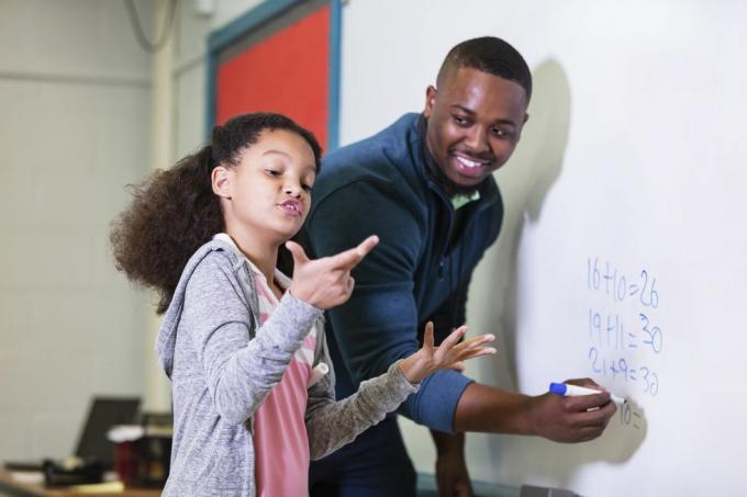 en multirasial 9 år gammel jente i matematikkklassen, står ved tavlen med læreren sin, en ung afroamerikansk mann i sin 20-årene ser læreren på grunnskoleeleven sin og venter mens hun prøver å finne ut svaret fokuset er på pike