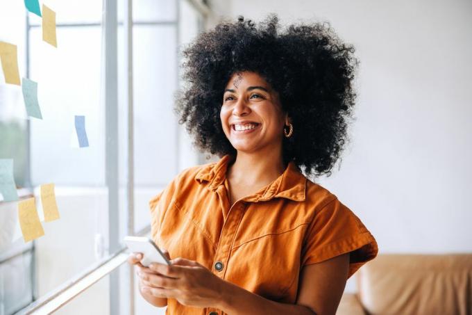 glad svart forretningskvinne smiler mens hun bruker en smarttelefon på et kreativt kontor munter kvinnelig gründer som sender en tekstmelding mens hun står ved siden av en glassvegg med klistrelapper