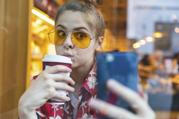 ung kvinne ute i solbriller og tar en selfie mens hun drikker en kaffe