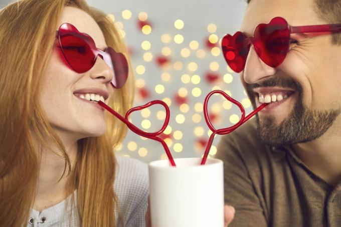 nærbilde portrett glad ung mann og kvinne forelsket i rosa solbriller nipper til en drink fra en kopp gjennom hjerteformede sugerør, nyter et søtt morsomt par øyeblikk på en morsom date på valentinsdagen
