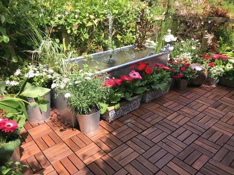 arkivbilde av hage trehus terrasse plattform balkong om sommeren med sinkmetall trau dam vann funksjon med solenergi fontene pumpe, gullfisk fisk, marginale planter, røde miniatyrroser, rosa gerbera blomster, teak terrassefliser, solcelledrevne lys, belysning